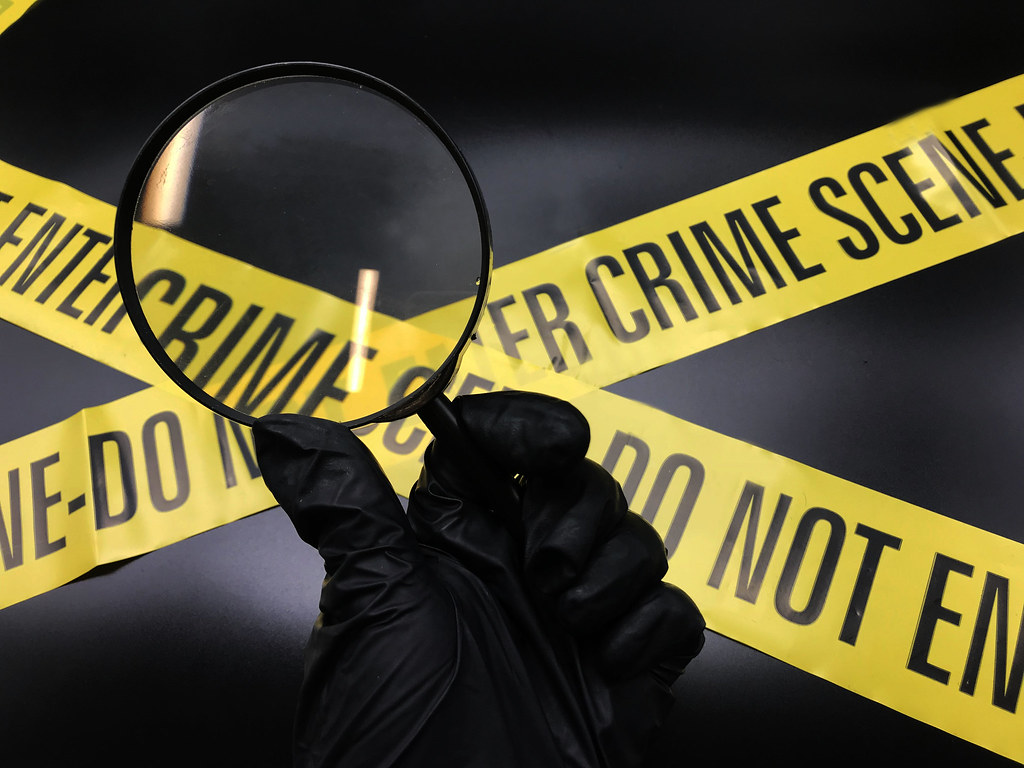 Yellow crime scene tape with a black gloved hand holding a magnifying glass over it